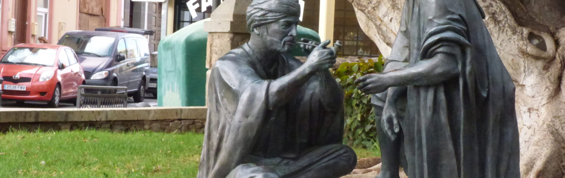 Two dark bronze statues in traditional dress seated on a decorative blue-and-white tilework base beneath a large tree in a town square.