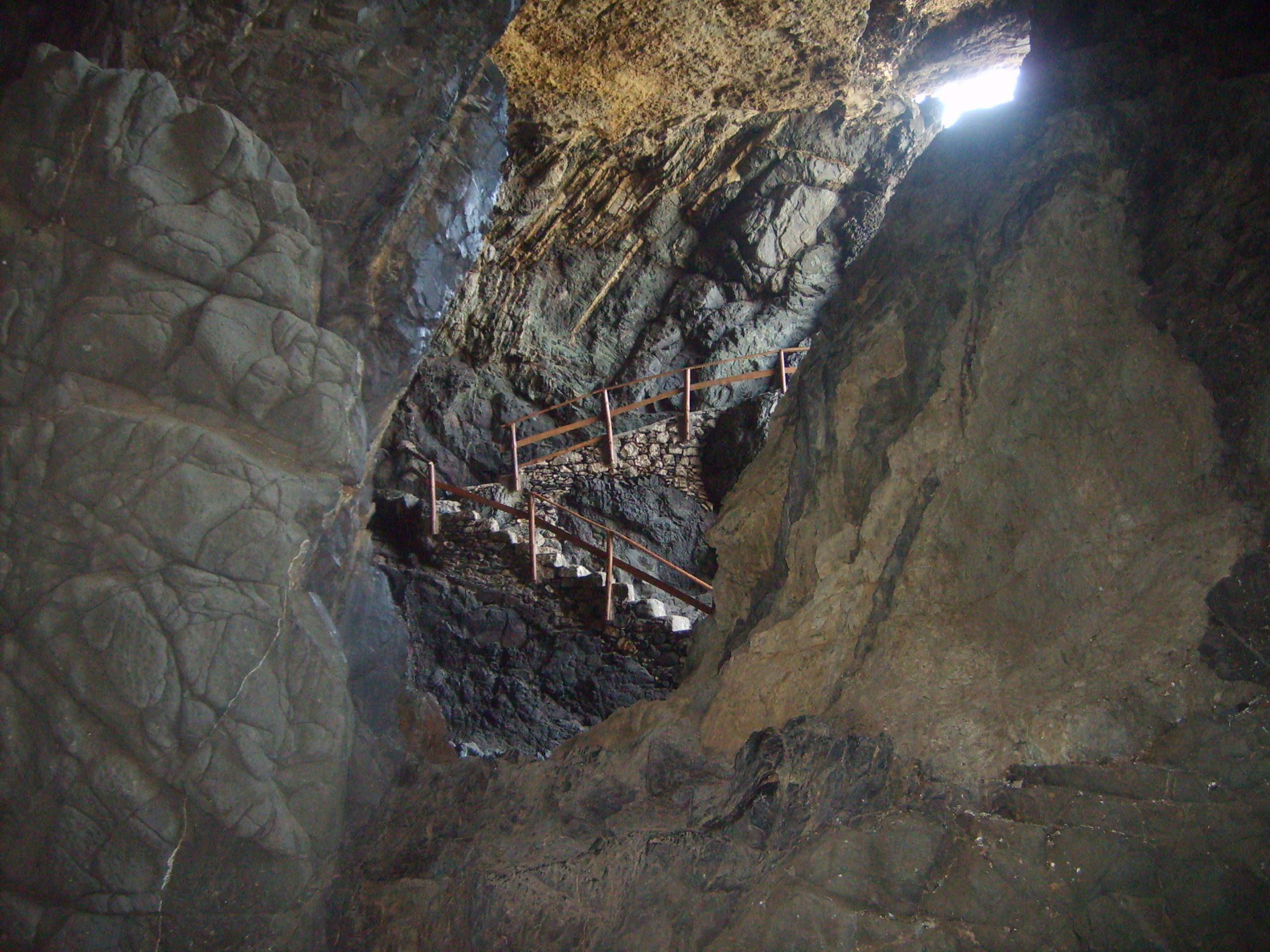 A metal stairway descends through the rocky passages of the Ajuy Caves, with natural light streaming in from the cave entrance.