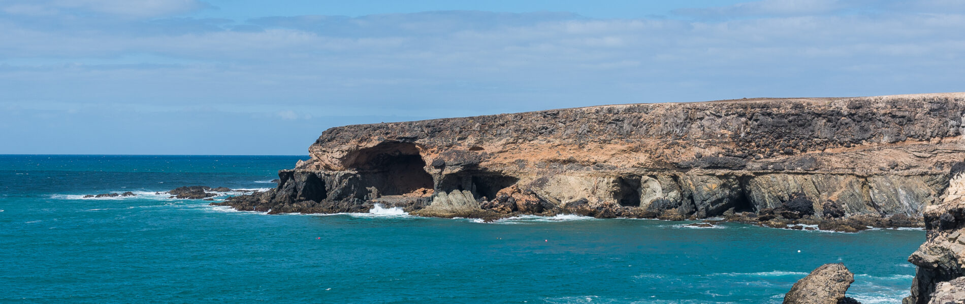 Rocky coastal cliffs with large dark cave openings overlooking turquoise Atlantic waters under clear blue sky