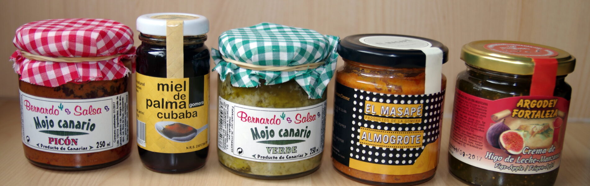 Five jars of Canary Islands food products lined up on a wooden surface, including mojo sauce, palm honey, and preserves with Spanish labels.