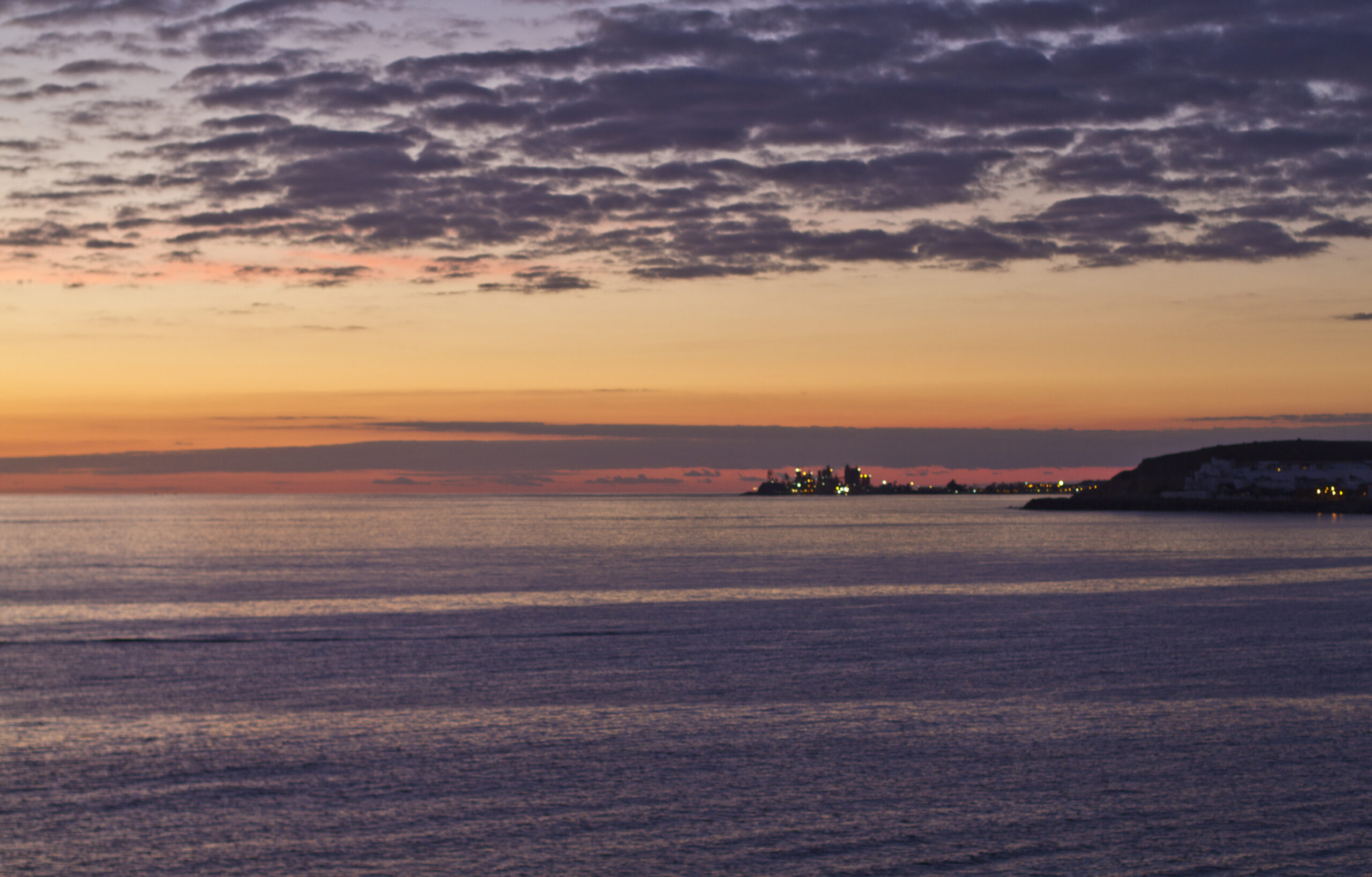 Arguineguín Beach at dusk, with the town's waterfront lights reflecting off the tranquil Atlantic waters under a dramatic twilight sky.