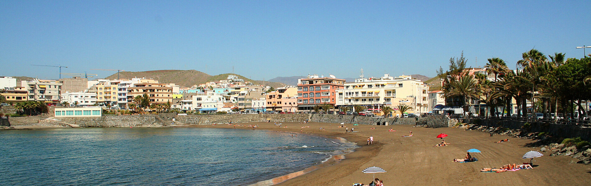 Sandy beach with turquoise water and scattered beachgoers, backed by a hillside town with colorful apartment buildings and palm trees under clear blue sky