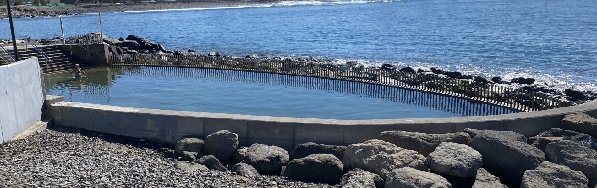 Natural tidal pool enclosed by wooden fencing with volcanic rocks and Atlantic horizon beyond.