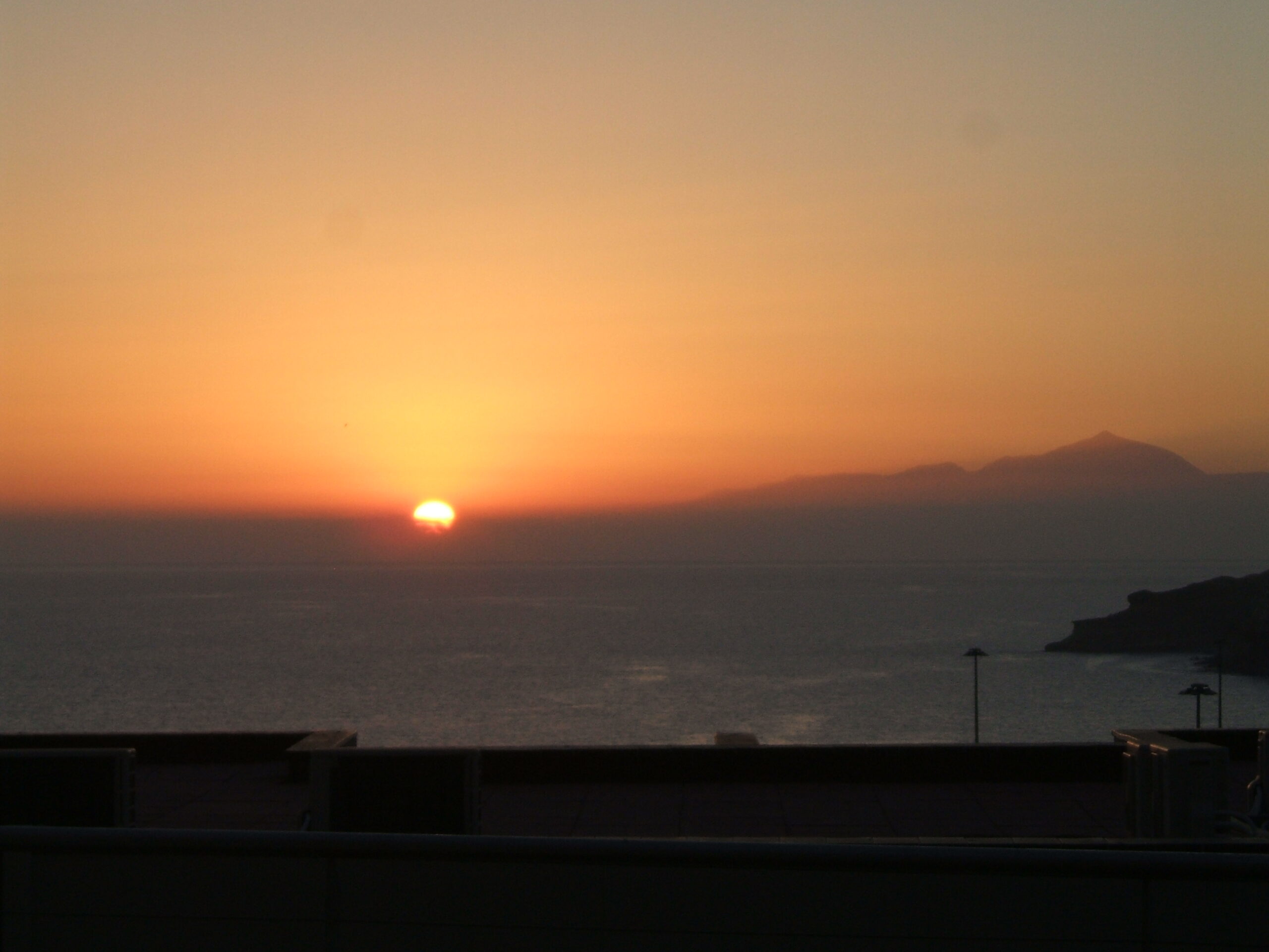 Warm sunset tones illuminate the Arguineguín coast, with a distant island silhouetted on the horizon.