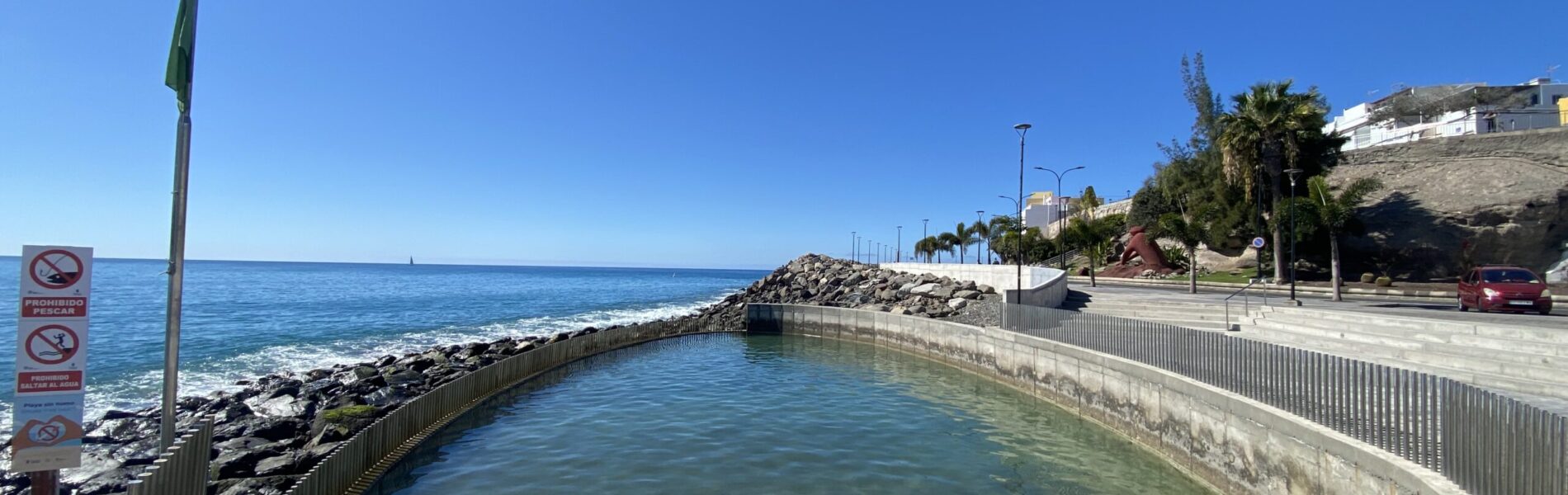 Sheltered harbour pool with turquoise water enclosed by rock jetties and coastal buildings.