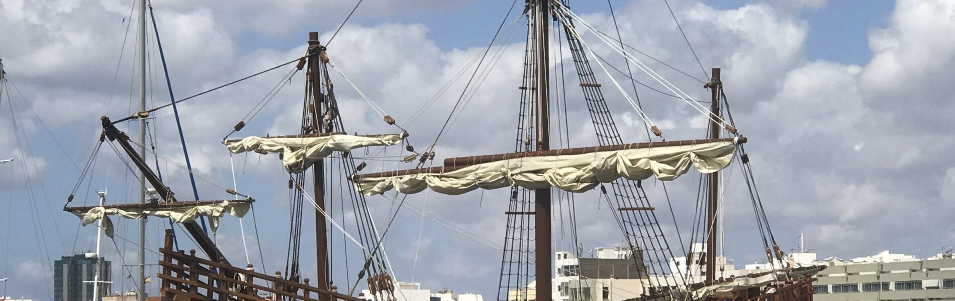 Replica caravel with dark wooden hull and cream-colored sails moored at a modern marina dock, with apartment buildings visible behind.