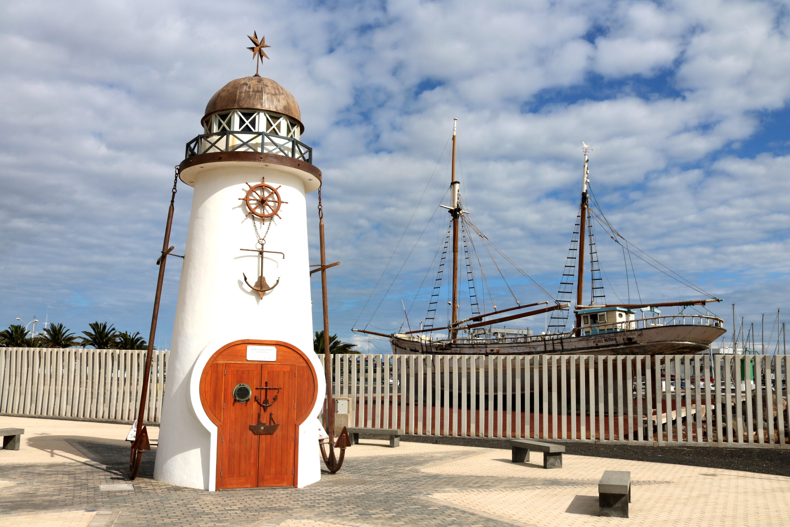 A charming decorative lighthouse with maritime details stands in the Arrecife Marina, accompanied by a traditional tall ship.