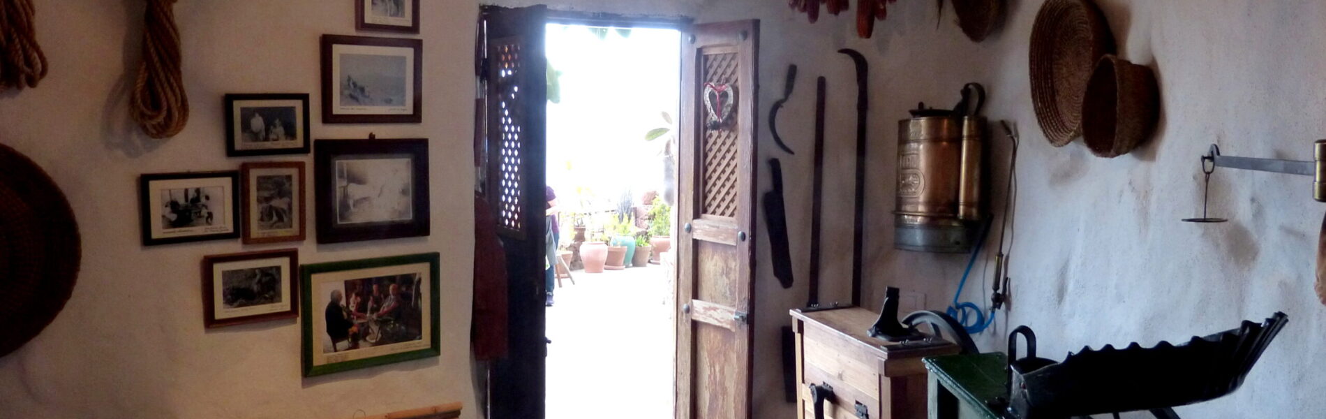 Interior of a whitewashed cave dwelling with dried peppers hanging from ceiling, framed photographs on walls, wooden furniture, traditional tools, and open doorway with bright daylight