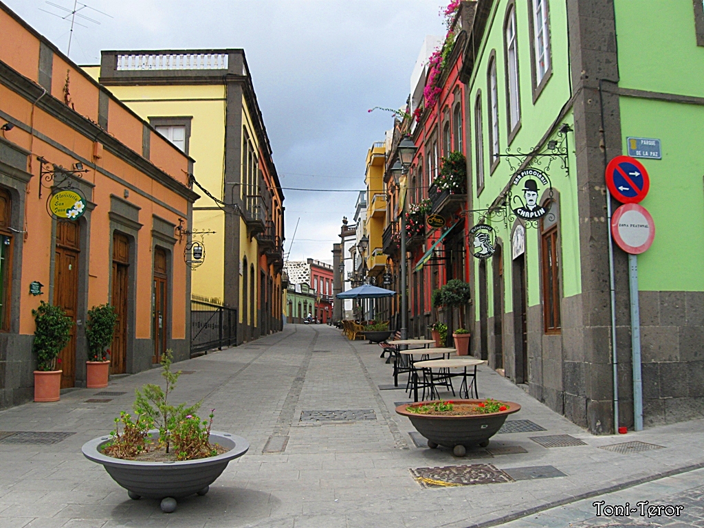 A vibrant pedestrian street in Arucas lined with historic colonial architecture painted in warm yellows, oranges, and greens, with traditional wrought-iron signs and flowering cascades adorning the facades.