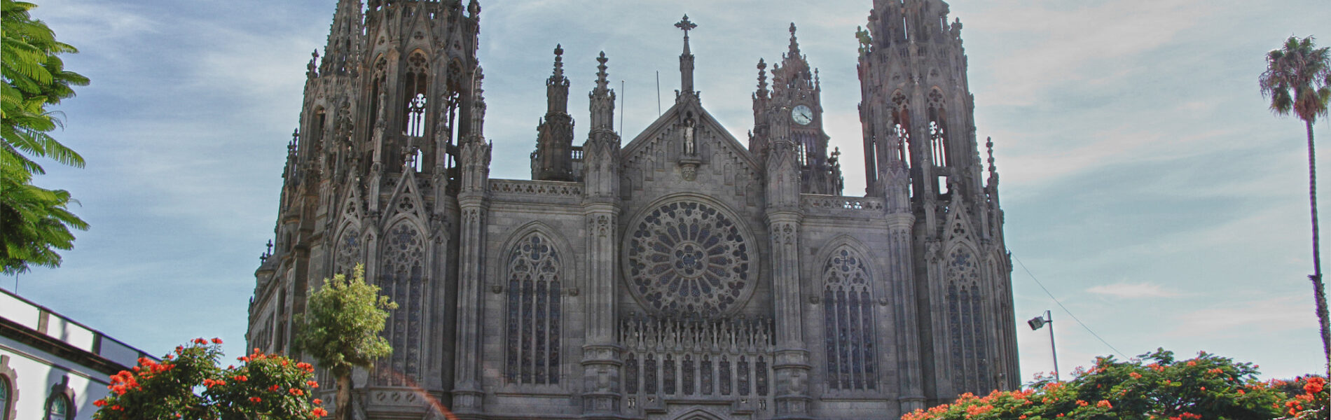 Neo-Gothic cathedral with twin spires, ornate stone facade, large rose window, and wooden doors, flanked by flowering shrubs and palm trees under partly cloudy sky.