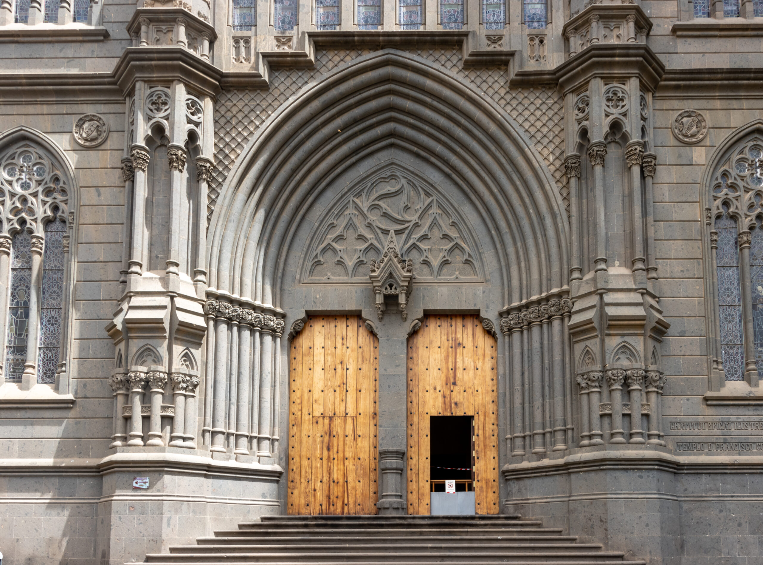 The imposing main entrance of Arucas Cathedral features a striking Gothic pointed arch framed by slender columns and intricate stone detailing.