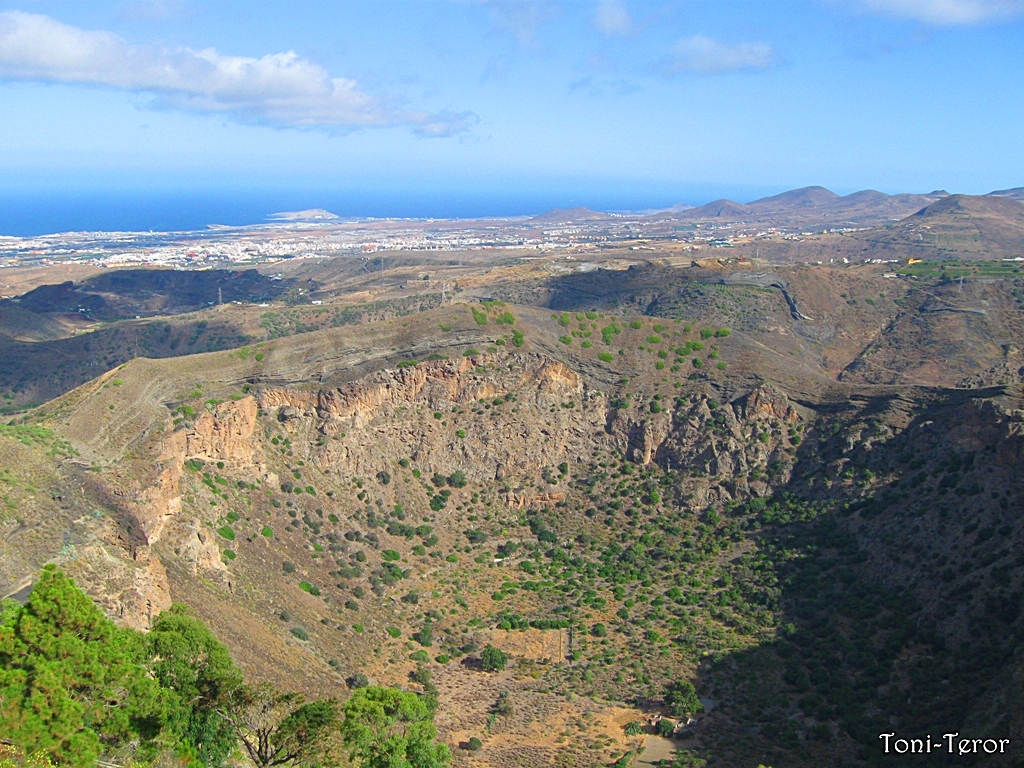 The dramatic crater of Bandama volcano reveals layers of volcanic rock and lava, with the northern coast of Gran Canaria visible in the distance.