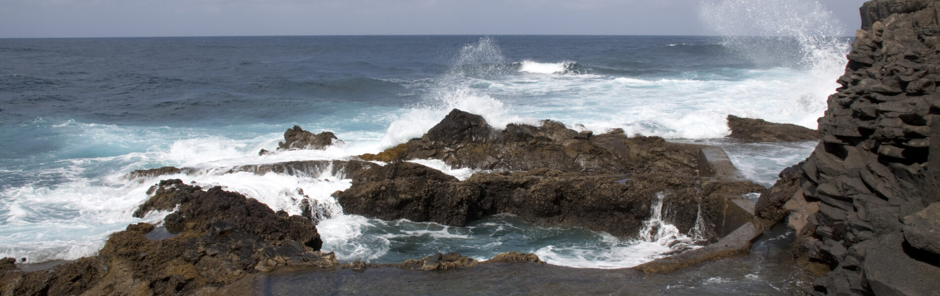 Rocky coastline with basalt formations, white waves crashing against dark rocks, natural tidal pool in foreground, Atlantic Ocean under partly cloudy sky