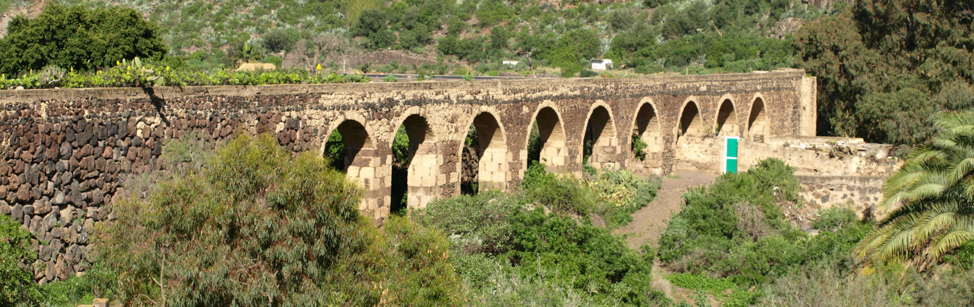 Stone aqueduct with multiple arches spanning a ravine, surrounded by vegetation and a rocky hillside