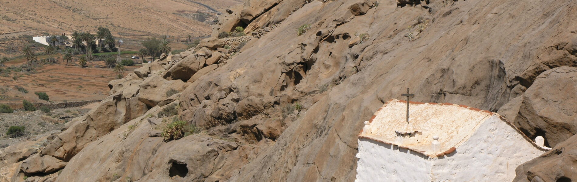 White-washed hermitage built into tan rocky hillside with sparse vegetation and barren mountains in background