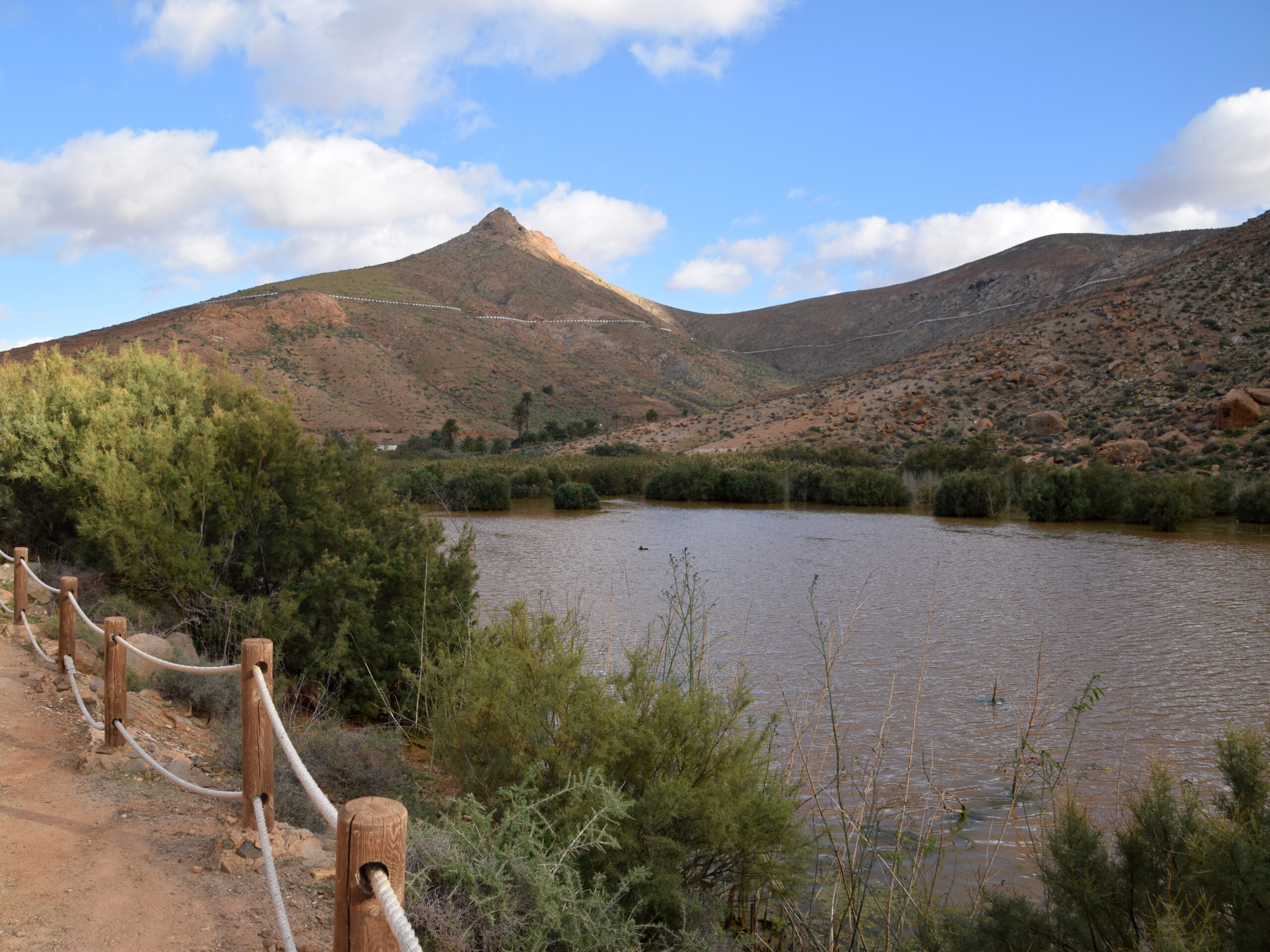 A distinctive volcanic peak rises above the tranquil waters of the reservoir at Barranco de las Peñitas, surrounded by native vegetation and volcanic terrain.