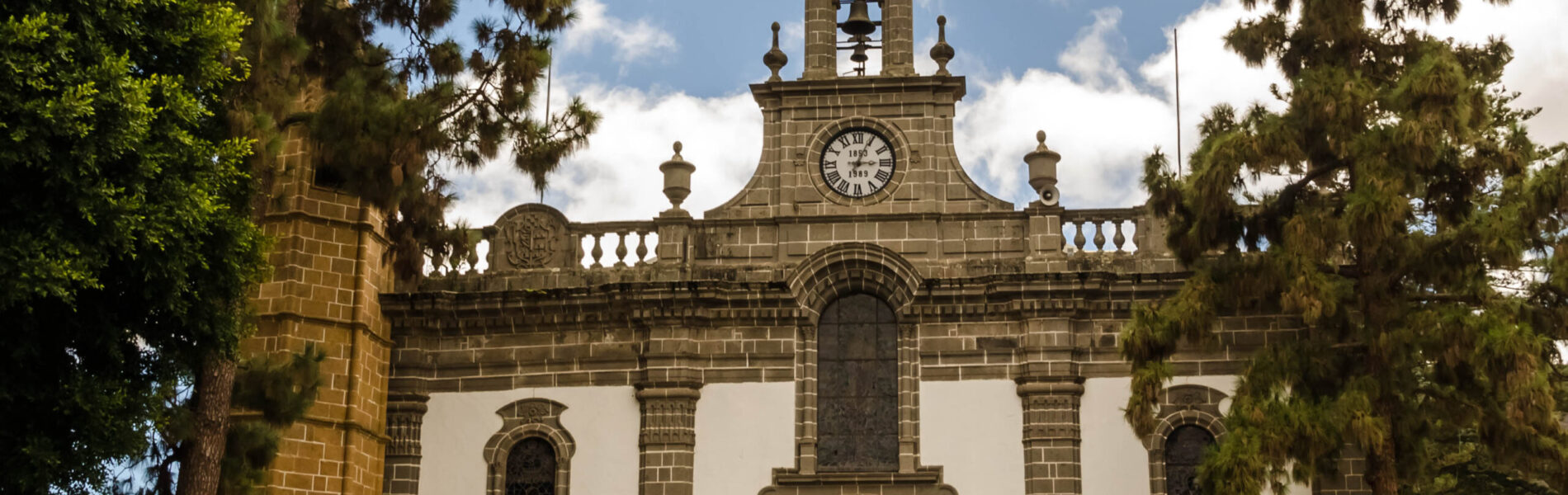 White and dark stone colonial church with clock tower framed by tall pine trees.