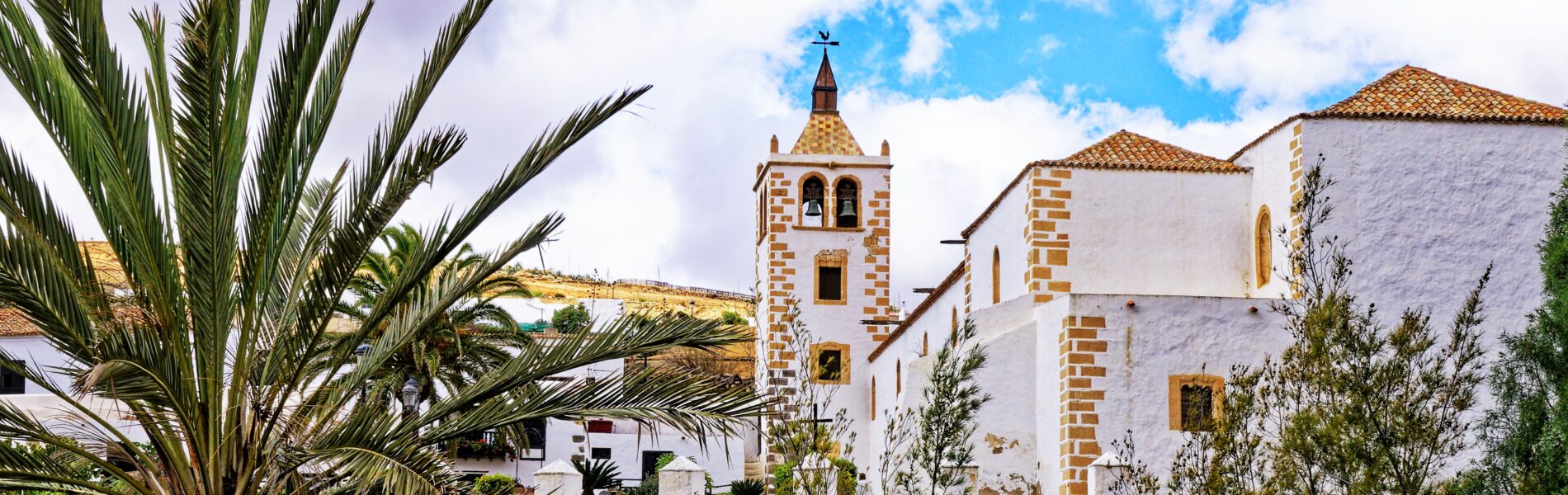 White-walled church with striped stone bell tower and terracotta roof, palm tree in foreground, hillside settlement behind