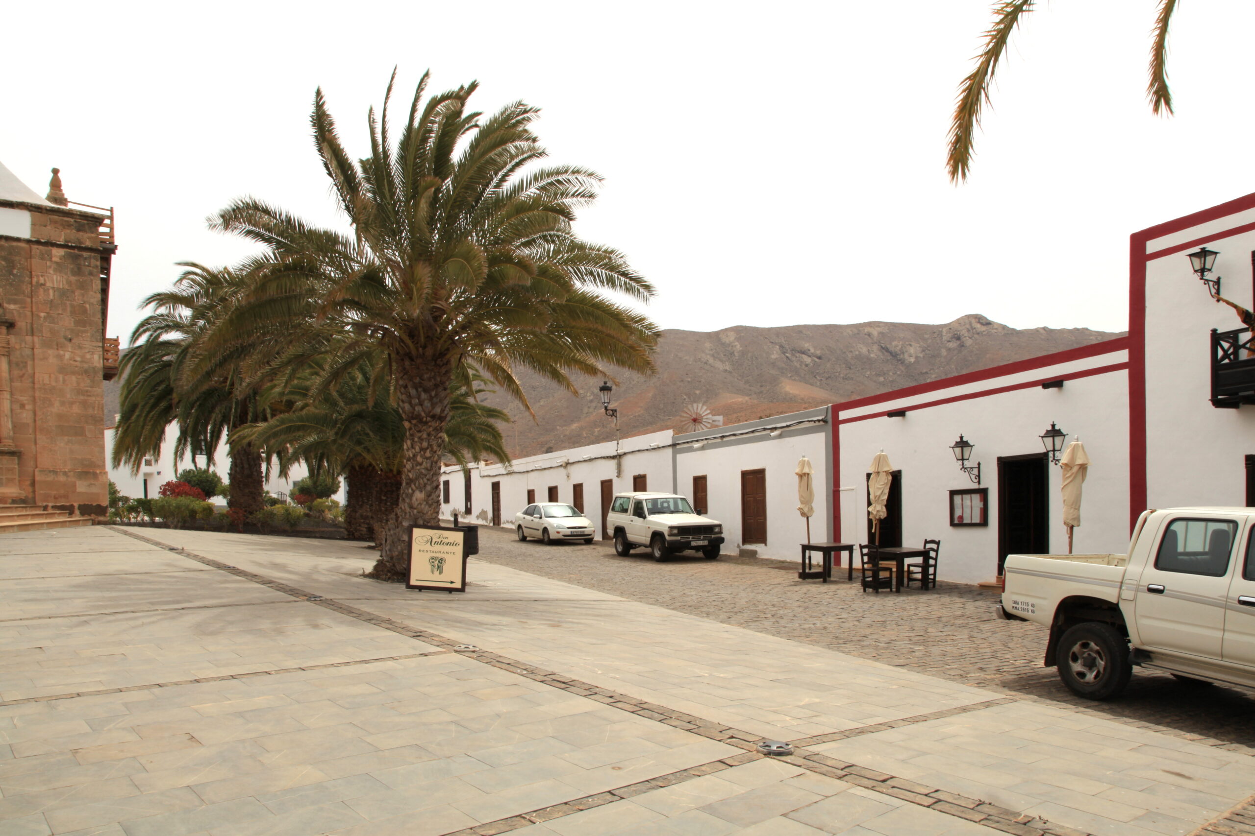The main plaza of Betancuria Old Town features traditional Canarian architecture with characteristic white walls and red accents, shaded by mature palm trees against the island's barren landscape.