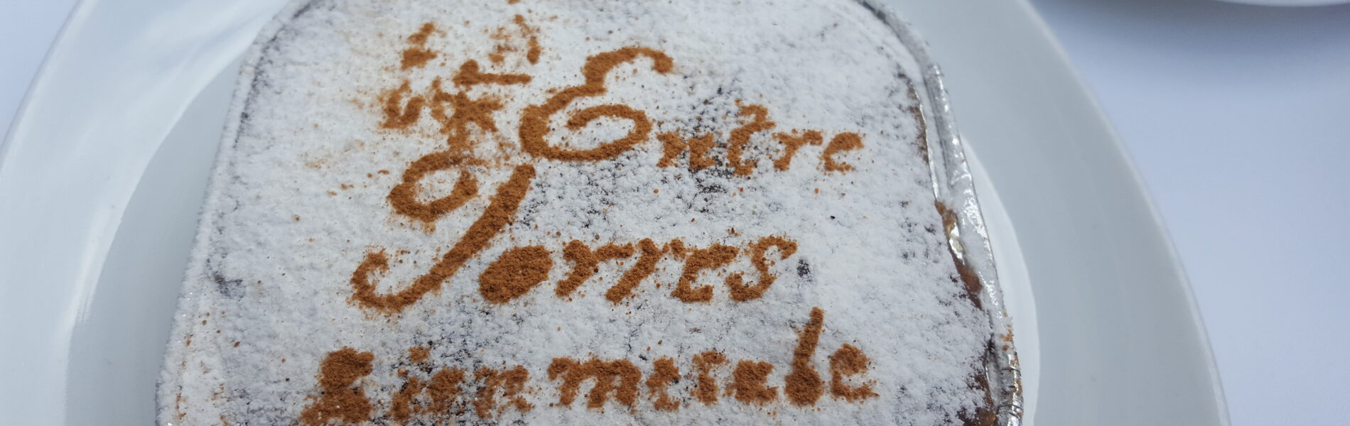 Square cake dusted with powdered sugar on a white plate, with text piped in cinnamon on top