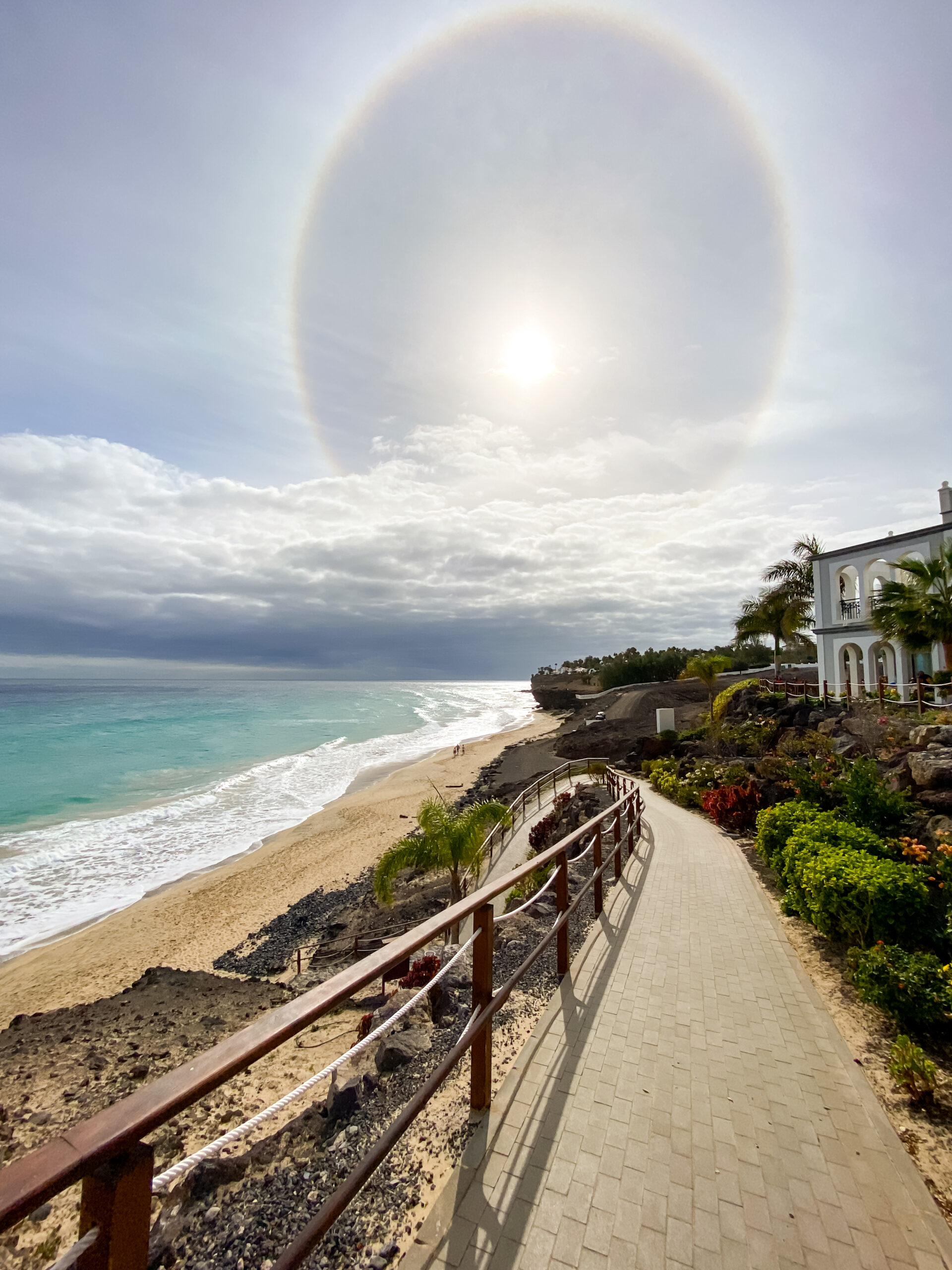 A paved waterfront path descends toward Butihondo Beach, where white-sand shores meet clear turquoise waters beneath a dramatic halo effect in the sky.