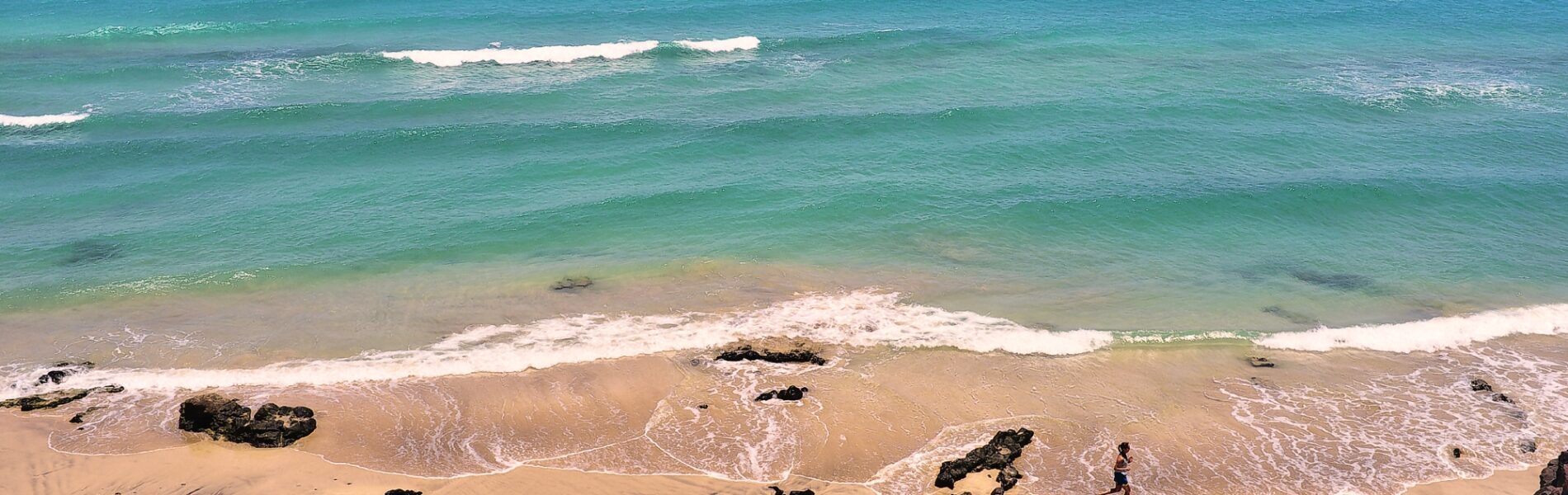 Sandy beach with turquoise water, white-capped waves, and dark volcanic rocks scattered along the shore under a partly cloudy sky.