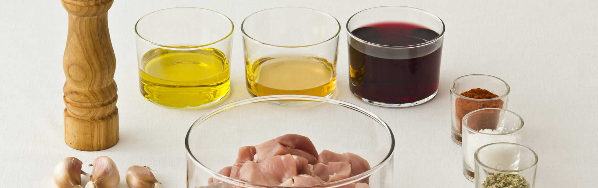 Wooden pepper mill, raw meat pieces in a bowl, olive oil, vinegar, wine, garlic, bay leaves, and spices arranged on white background.