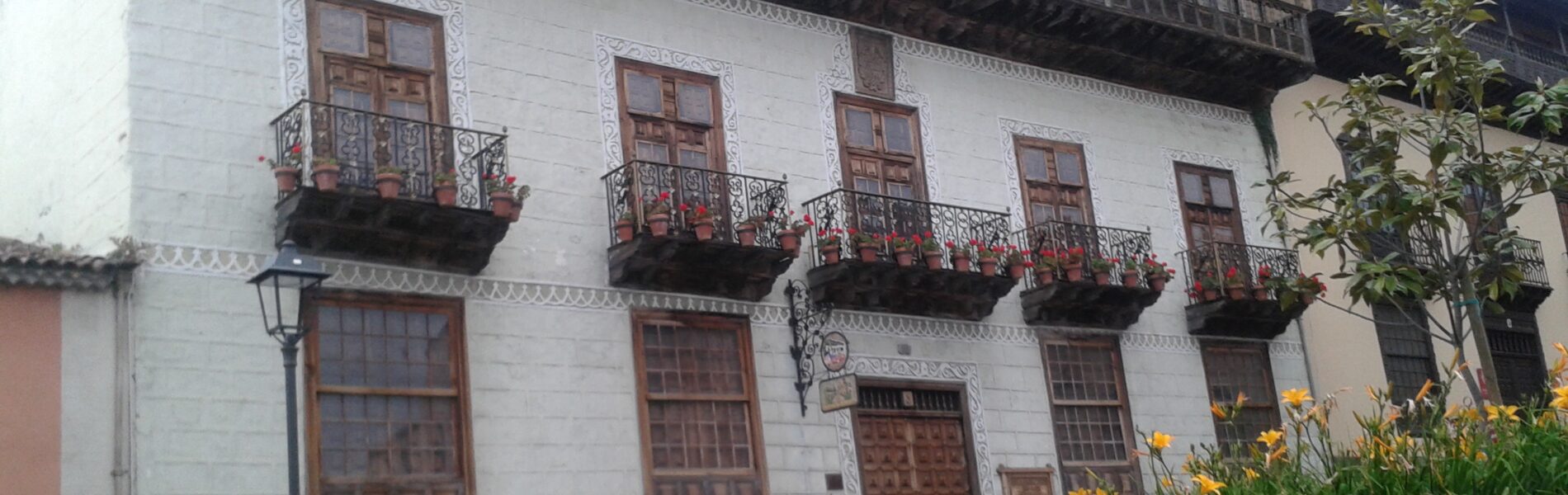 White colonial building with dark wooden balconies and ornate carved window frames, featuring a darker stone base and traditional architecture.
