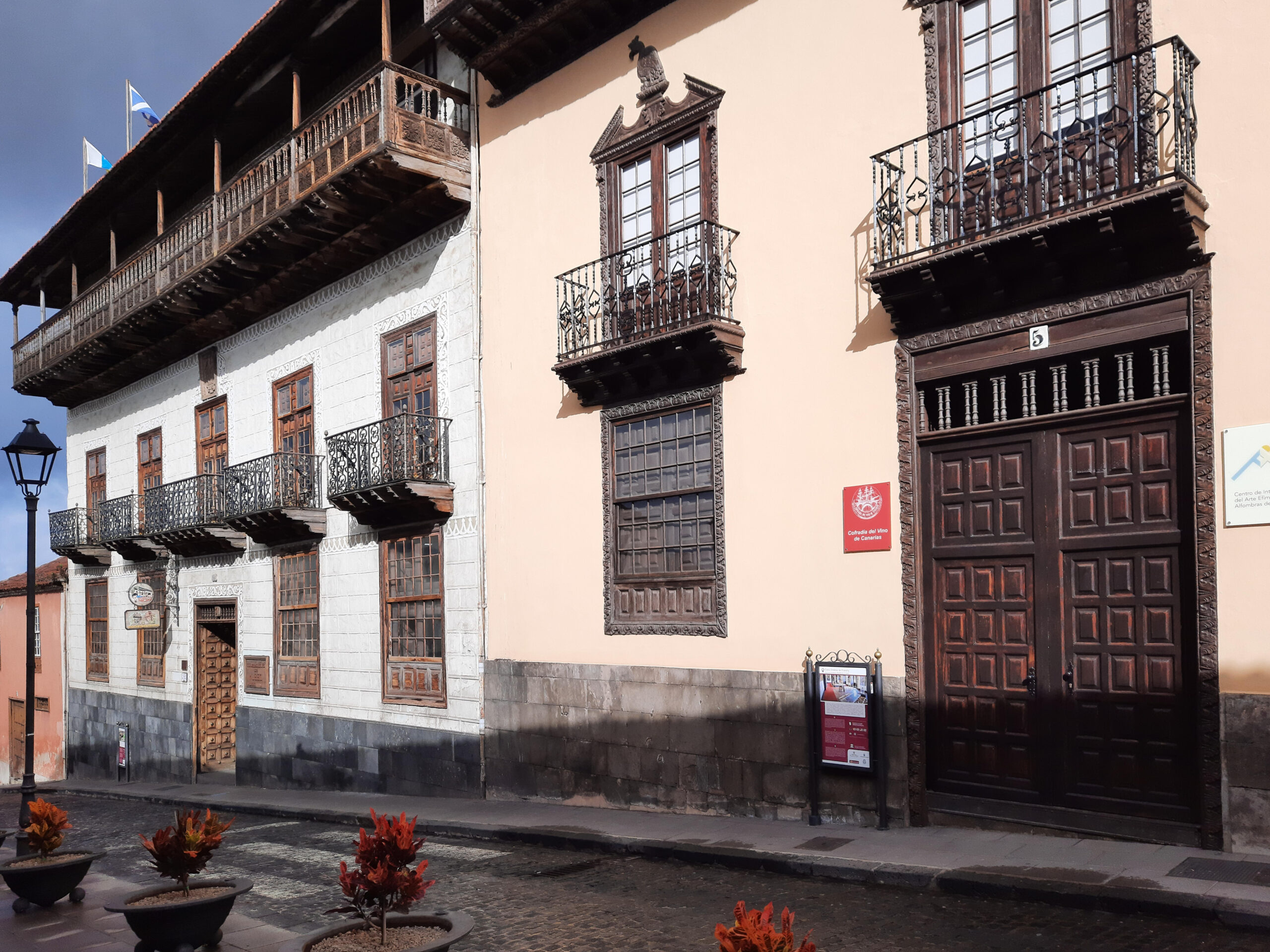 Colonial-era whitewashed building with dark wooden carved balconies, ornate windows, and a large burgundy door on a cobblestone plaza.