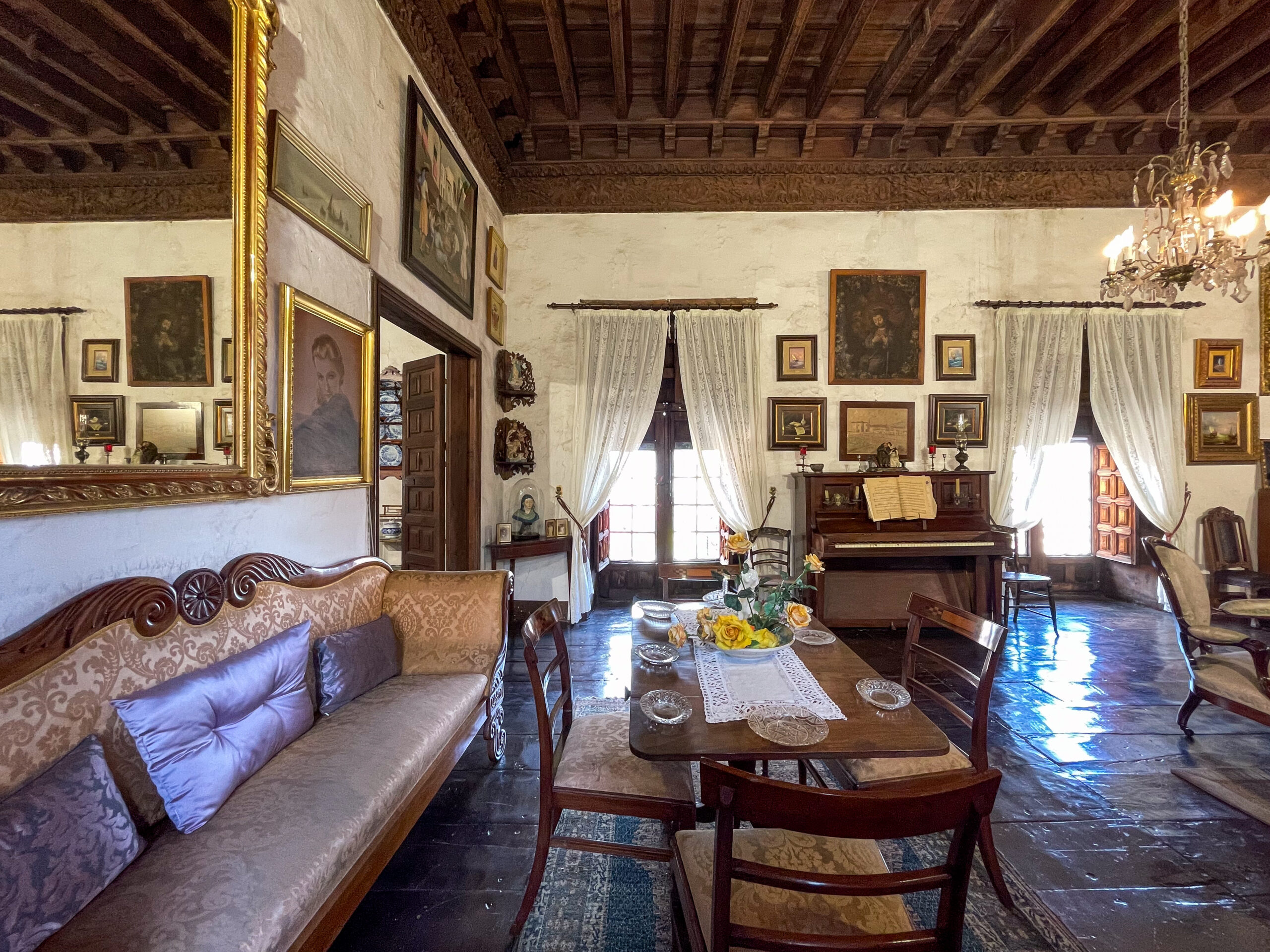 Historic interior room with ornate wooden ceiling, cream walls lined with framed paintings, period furniture including carved sofa and piano, chandelier, and sheer curtains framing tall windows.