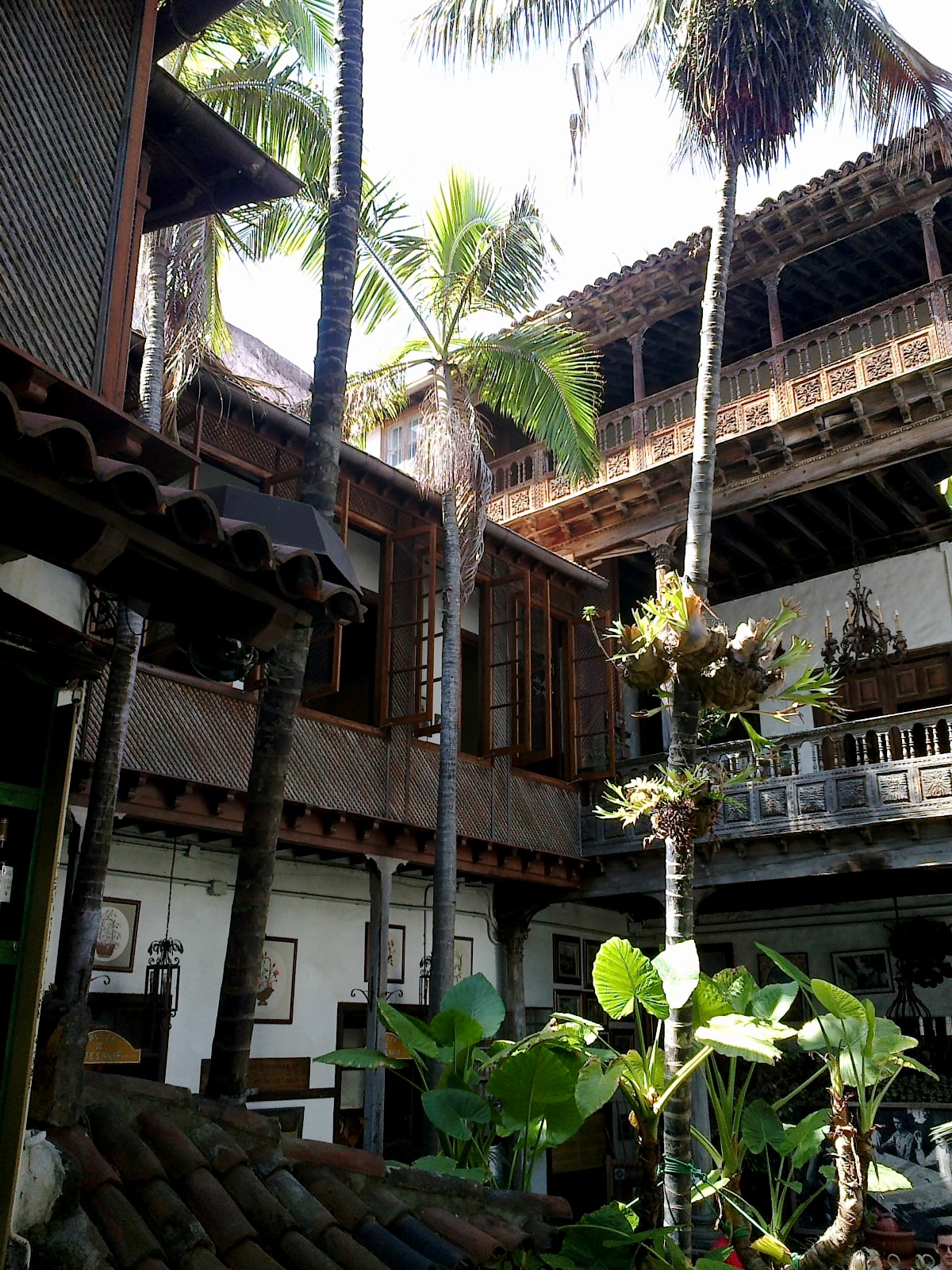 Wooden colonial courtyard with carved balconies, tile roofs, palm trees, and white-walled arcades around a central open space.