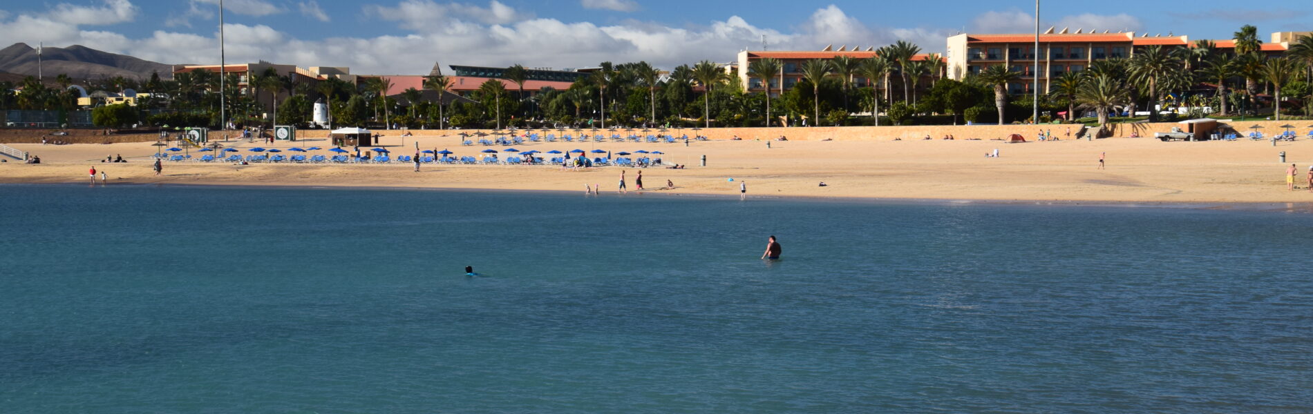 Sandy beach with clear turquoise water, blue umbrellas, palm trees, and a white building with orange roof under blue sky