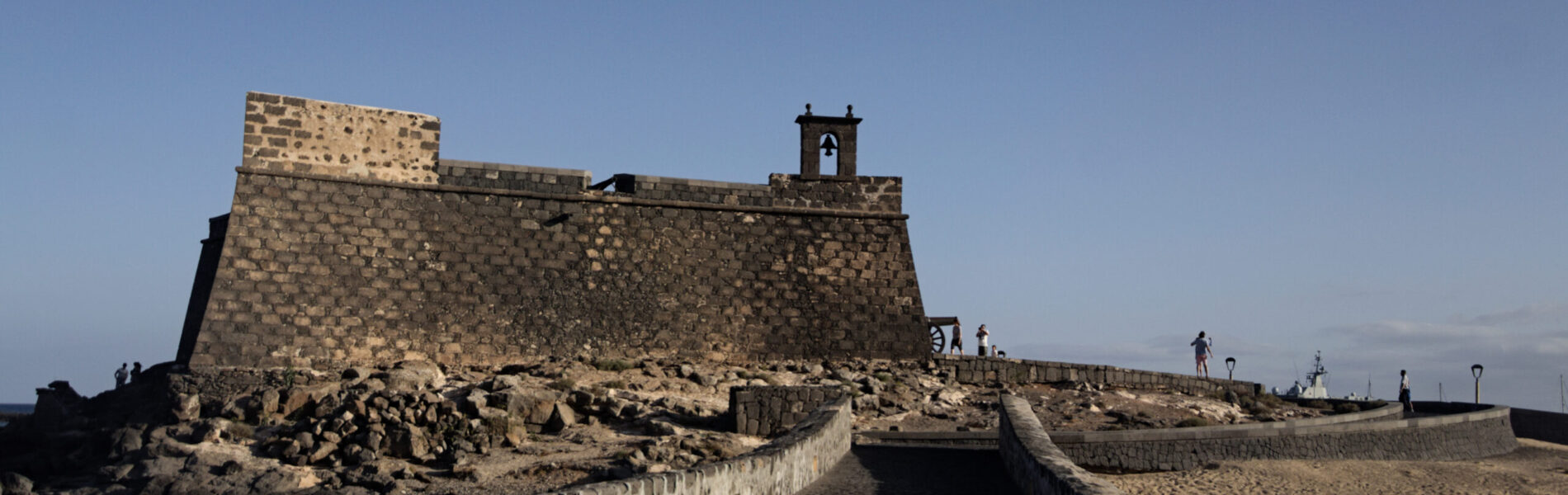 Stone fortress with crenellated walls and a small bell tower, accessed by a causeway across dark volcanic rock and sand under clear blue sky.