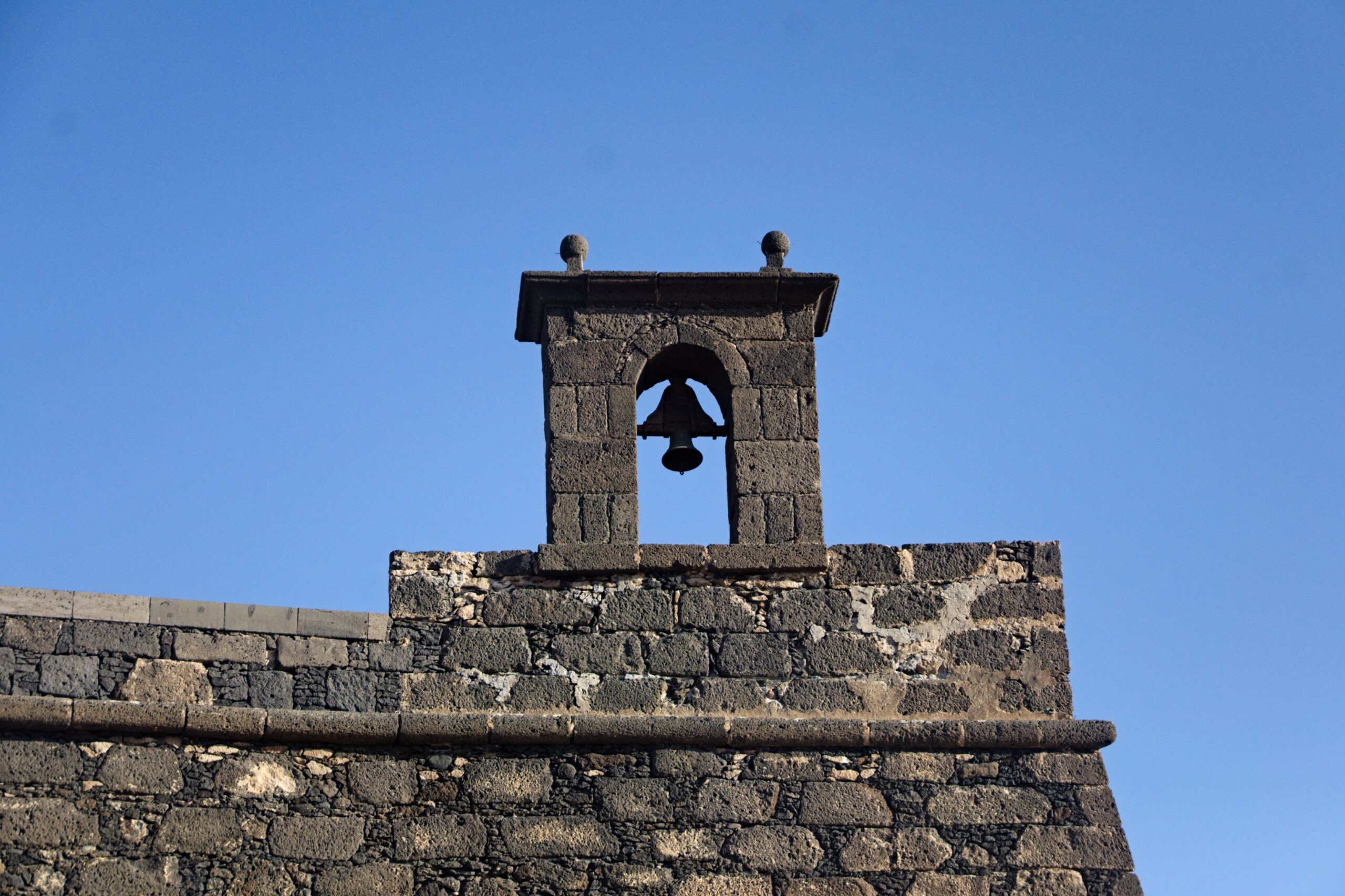 The bell tower of Castillo de San Gabriel overlooks the harbor of Arrecife with its distinctive dark bell framed against the sky.