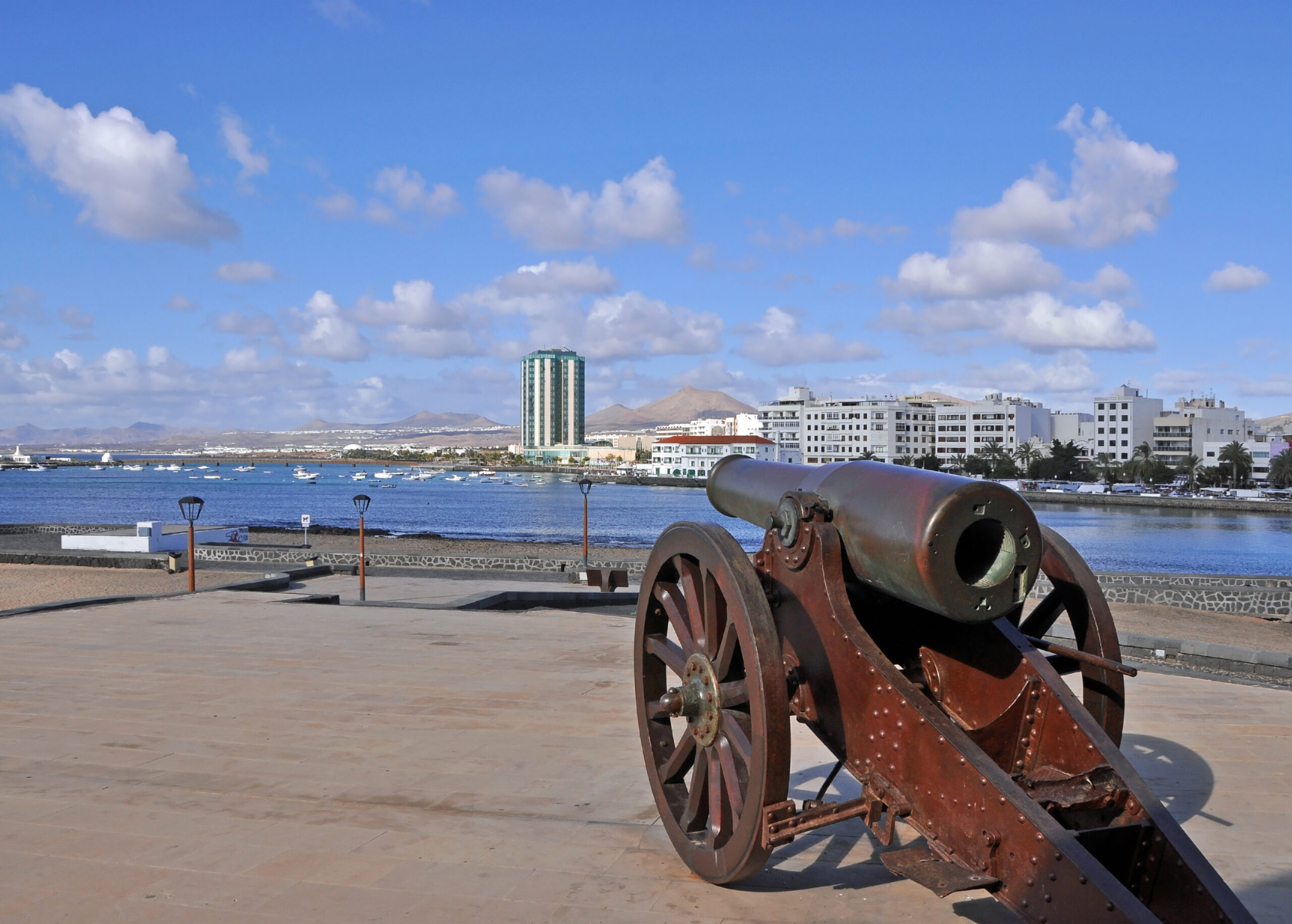 A historic cannon on the Arrecife waterfront commands a view across the harbor toward the distinctive modern architecture and volcanic peaks of Lanzarote.