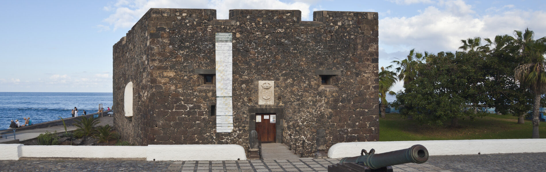 Stone fortress with crenellated walls and a central whitewashed stripe, facing the sea with a cannon in the foreground and palm trees nearby.