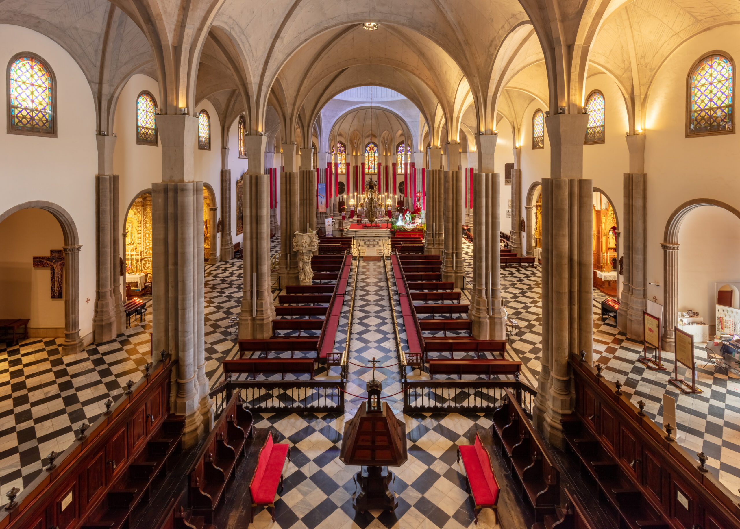 Interior of a Gothic cathedral with vaulted ceiling, stone columns, wooden pews, stained glass windows, and an altar framed by red decorative panels at the far end.