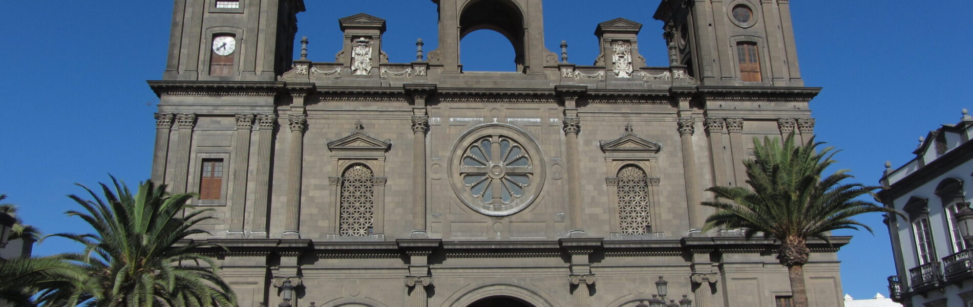 Basalt‑stone cathedral with twin towers, central dome, rose window and three arched entrances, framed by palm trees under a clear blue sky.