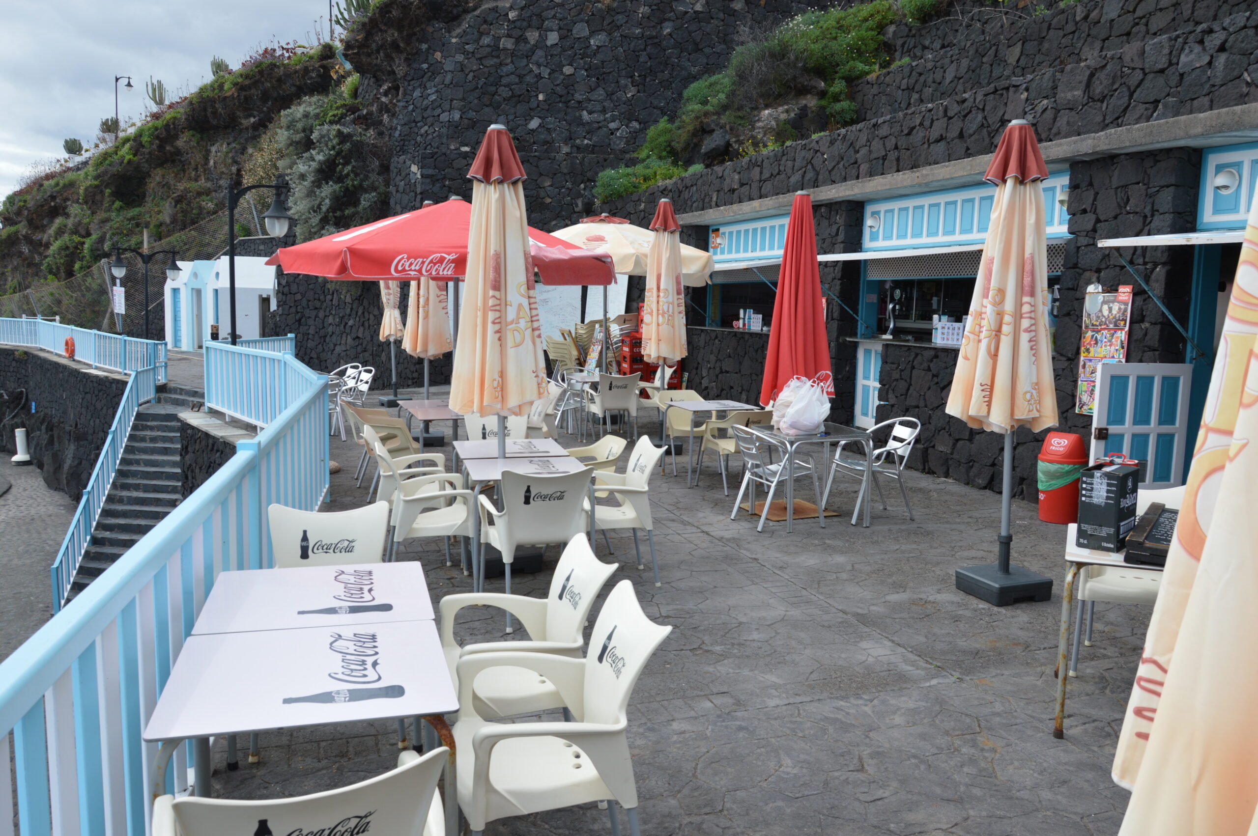 A seaside café at Charco Azul features traditional blue and white shuttered buildings built into the volcanic rock, with shaded seating beneath red and cream parasols.