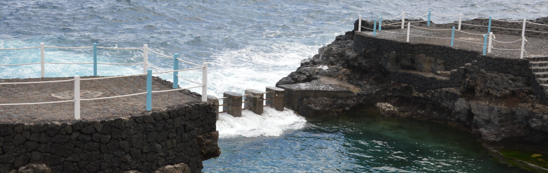Natural seawater pool surrounded by dark volcanic rock walls with white and blue railings, waves breaking beyond, ocean and sky visible.