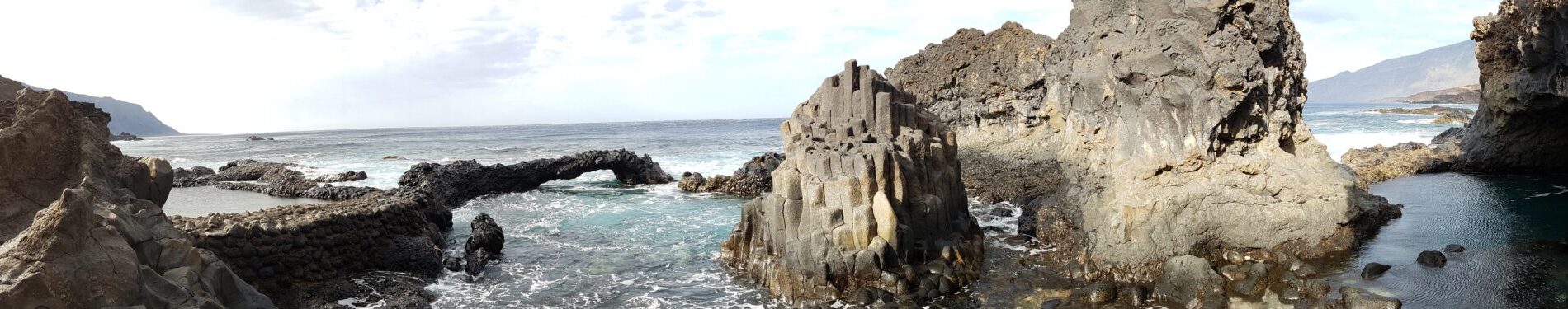 Rocky coastline with columnar basalt formations, turquoise pools, and ocean backdrop under cloudy sky