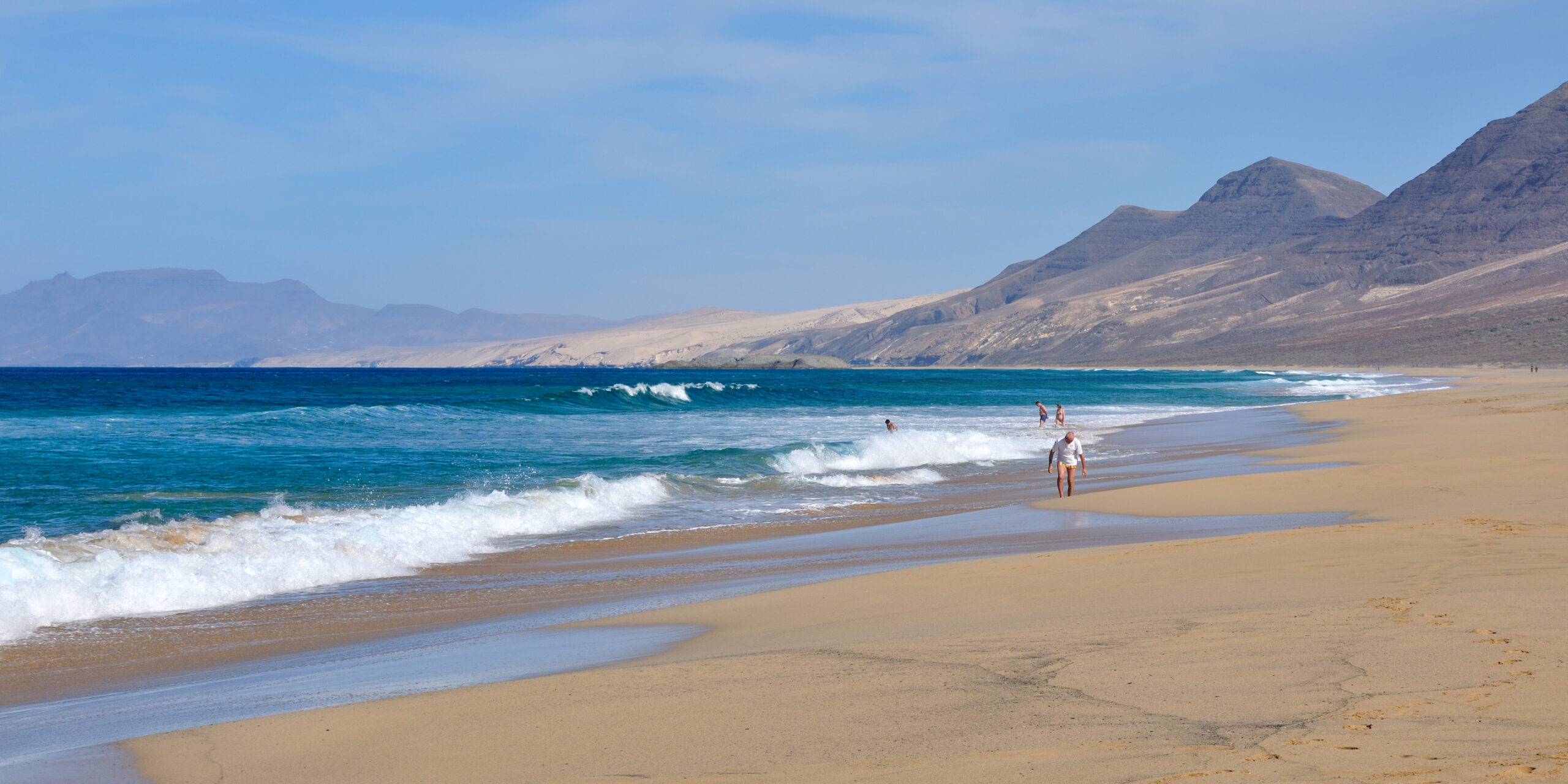 Cofete Beach stretches along Jandía's coast with golden sand, rolling Atlantic waves, and dramatic volcanic peaks rising behind the shoreline.