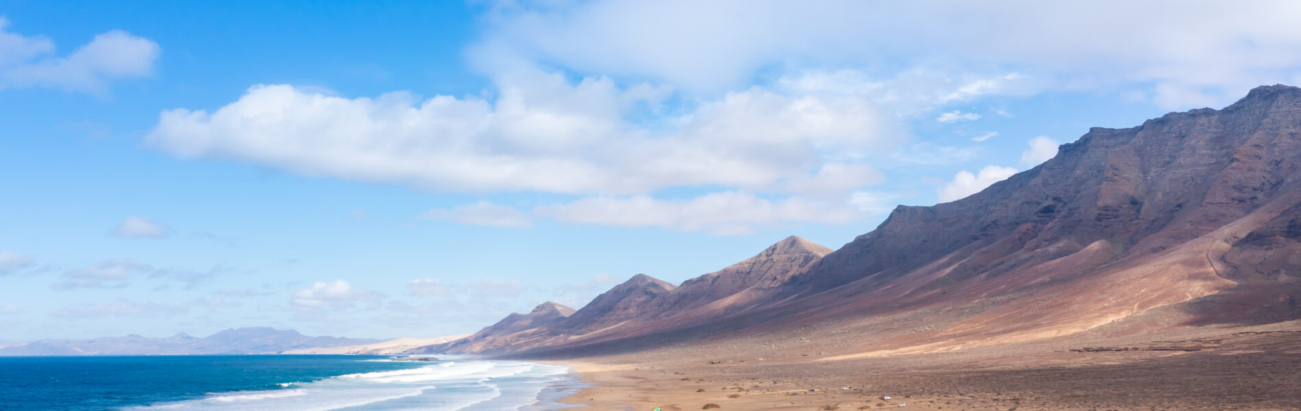 Wide sandy beach with breaking waves, backed by arid volcanic mountains under blue sky with white clouds