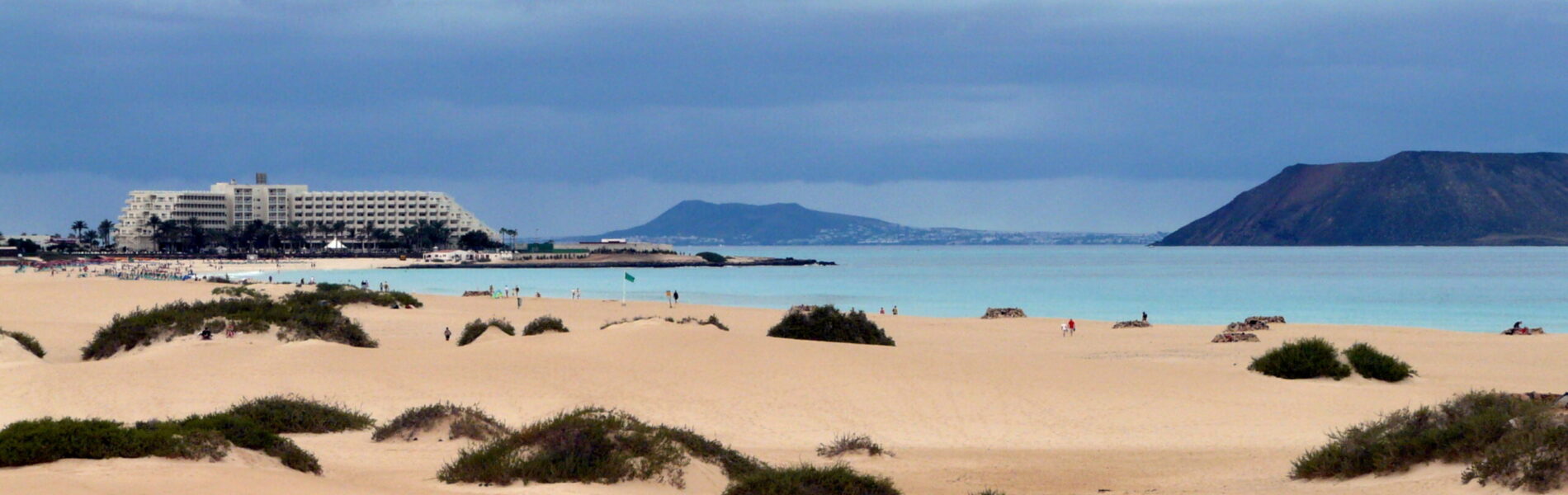 Wide sandy beach with sparse vegetation, turquoise water, beachfront hotel complex, and volcanic islands visible across the bay under cloudy sky
