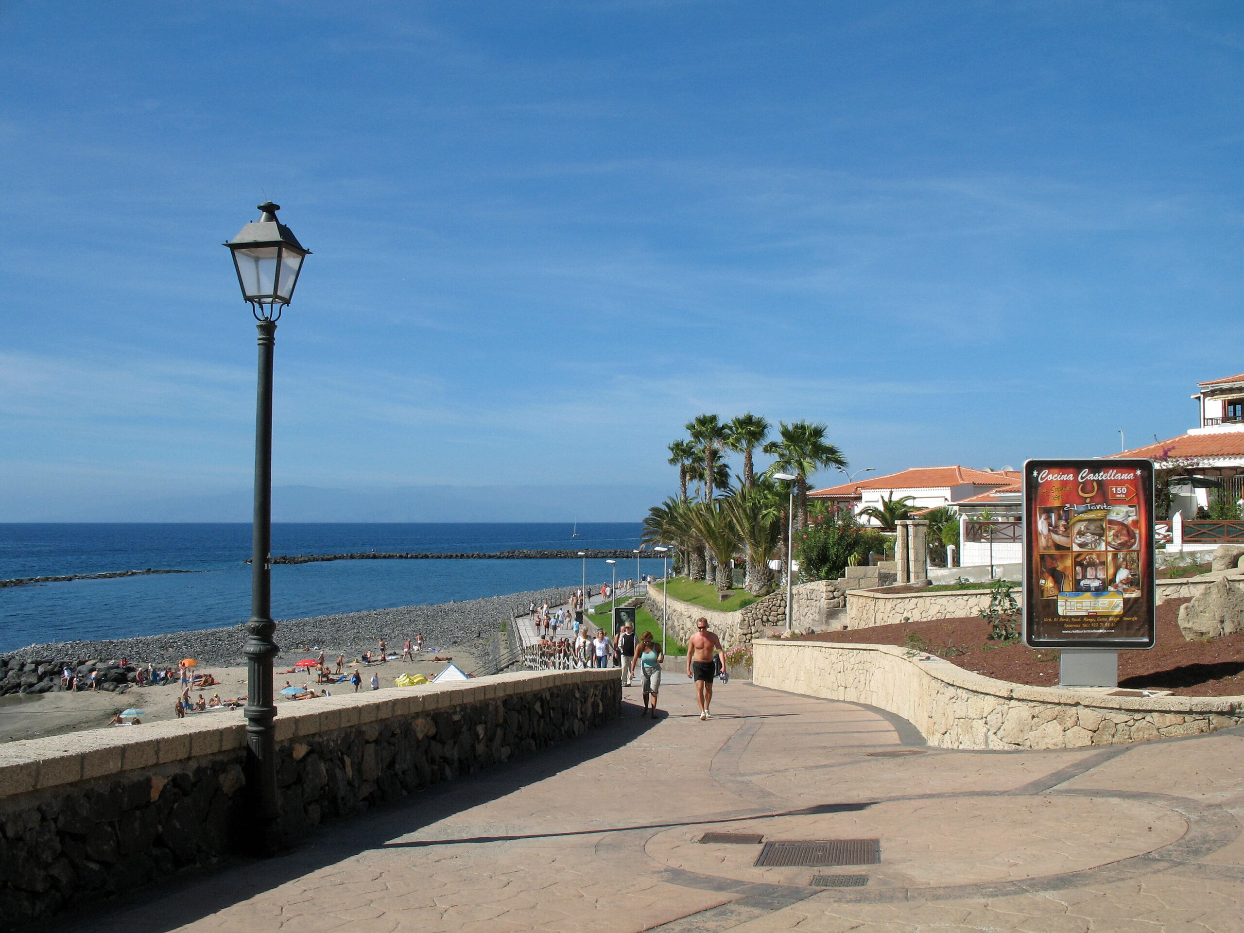 Beachside promenade with stone walls, lamp post, palm trees, and coastal buildings overlooking a sandy beach and blue ocean