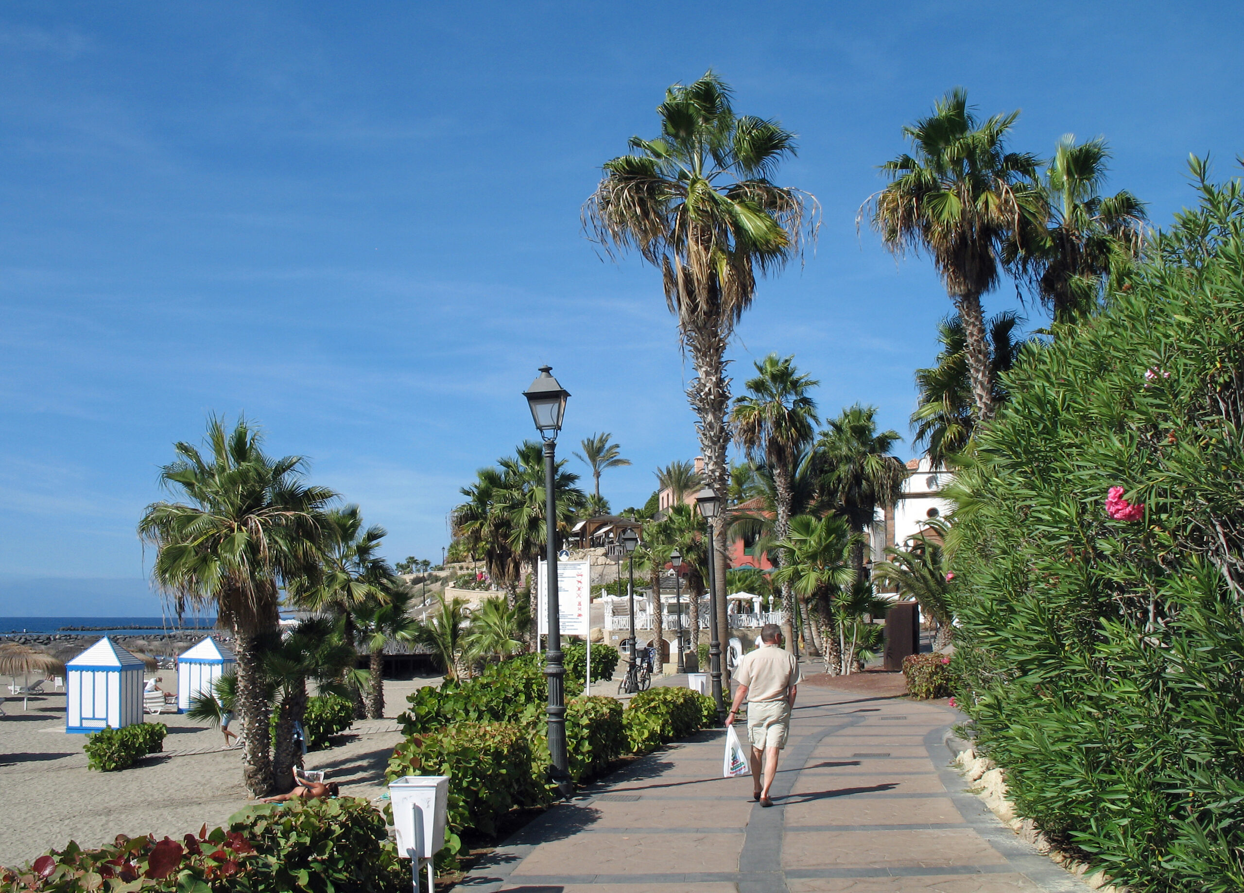 Beachfront promenade with palm trees, striped beach cabins, and white-walled buildings under clear blue sky