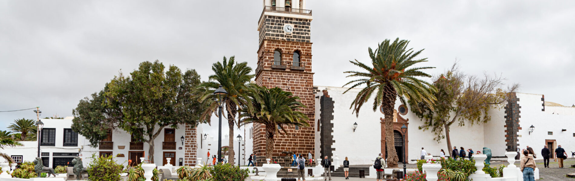 White-walled church with red stone bell tower and golden dome, flanked by palm trees in a cobblestone plaza with visitors.