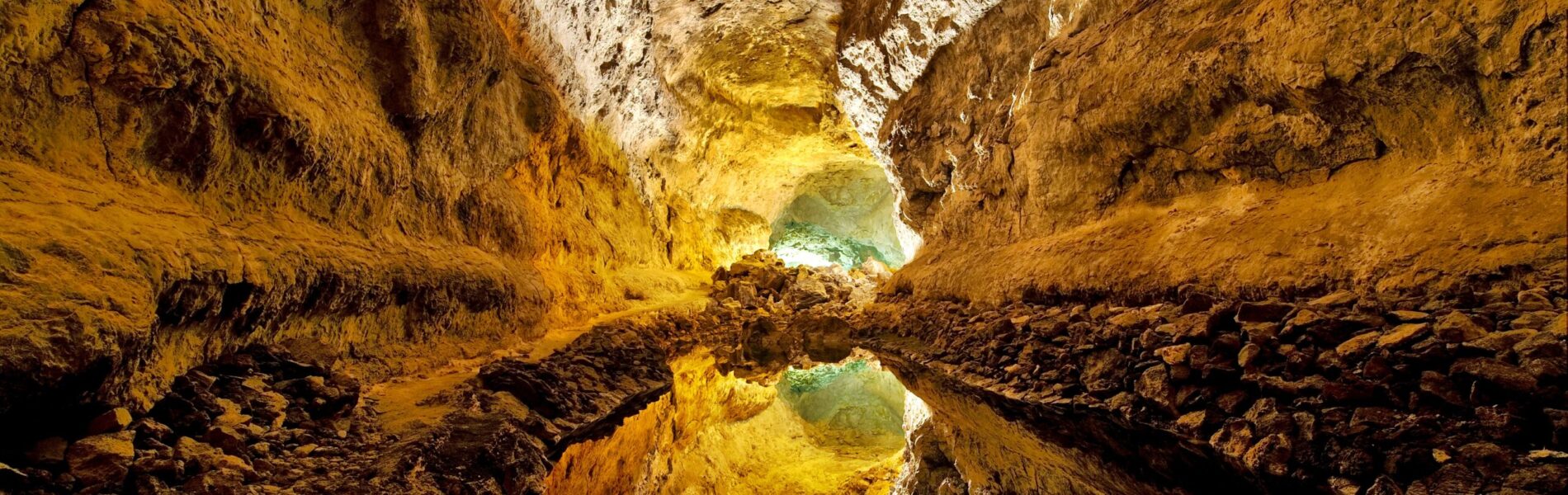 Underground cave tunnel with golden-yellow volcanic rock walls, rocky floor, and a pool of water reflecting sunlight streaming through an opening ahead.