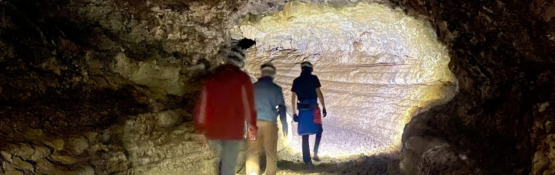 Three visitors walking through a large volcanic lava tube with rough stone walls, lit by natural light from an opening ahead.