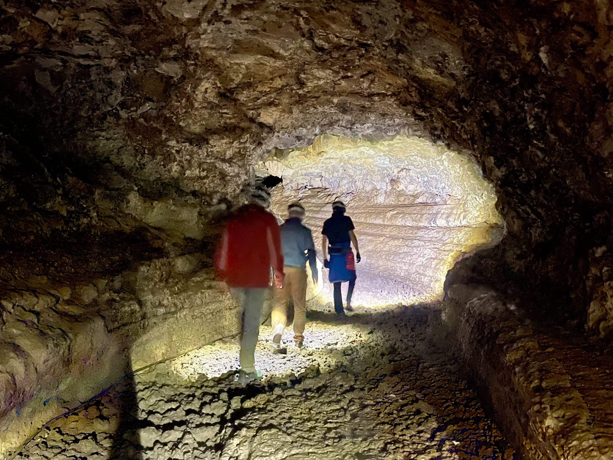 Three visitors walking through a large volcanic lava tube with rough stone walls, lit by natural light from an opening ahead.